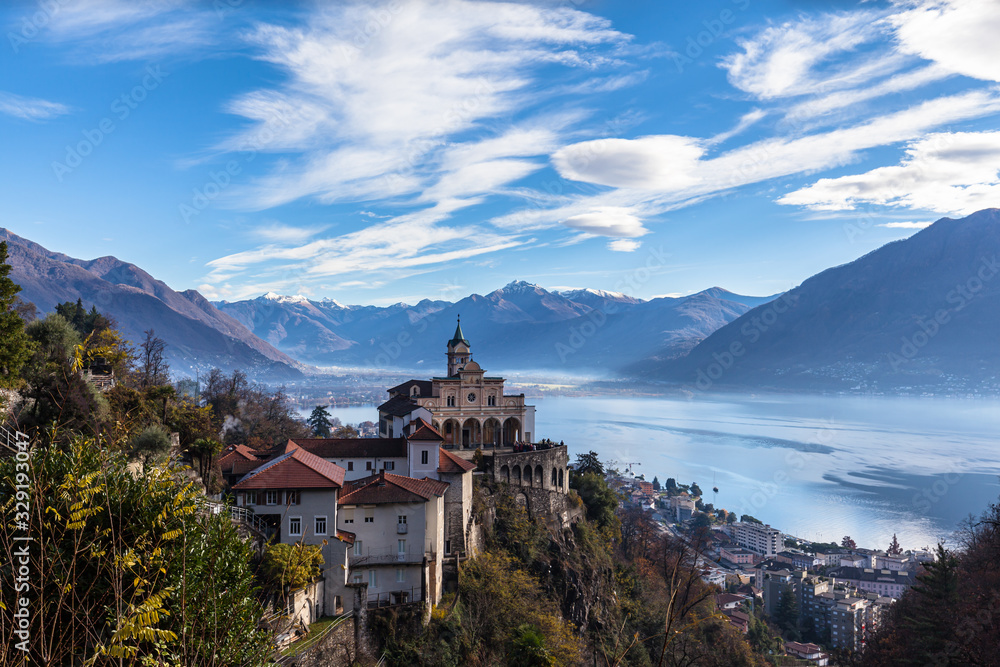 Stunning aerial panorama view of Madonna del Sasso church above Locarno city with Lake Maggiore, snow covered Swiss Alps mountain peaks and blue sky cloud in background in autumn, Ticino, Switzerland