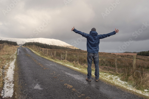 Man standing on a small road hand up in the air, road leads to a mountains covered with snow, concept outdoor, excitement, travel.