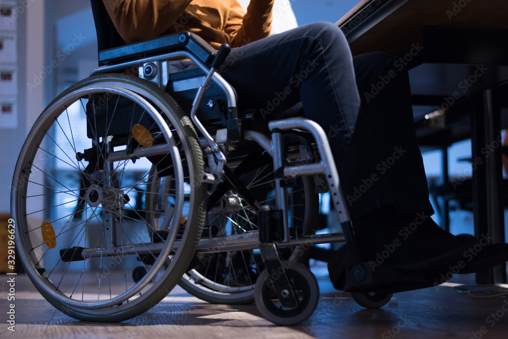 Disabled businessman in the wheelchair works in the office at the computer while performing in co-working space. Disability and handicap concept. Horizontal shot. Selective focus. Disabled businessman in the wheelchair works in the office at the computer while performing in co-working space. Disability and handicap concept. Horizontal shot. Selective focus.