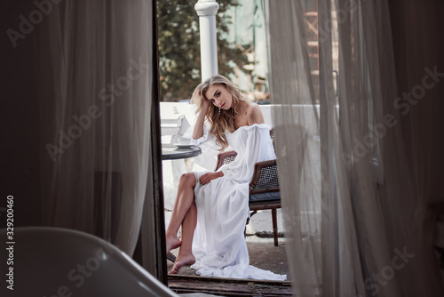 A tender bride in a light white dress drinks coffee on the outdoor terrace. Light and gentle image of the bride.
