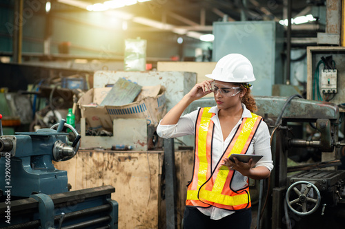 Female industrial engineer wearing a white helmet while standing in a heavy industrial factory behind she looking of working at industrial machinery and check security system setup in factory.