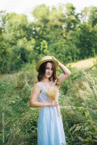 a cute and stylish girl in a white dress and long hair in the sunny summer park. A beautiful young girl in a white dress standing near the green tree