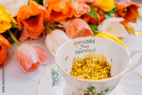 many Golden spangles of stars in white porcelain mug with text may month on a smooth light white background. Bouquet of multi-colored tulips on the background. photo