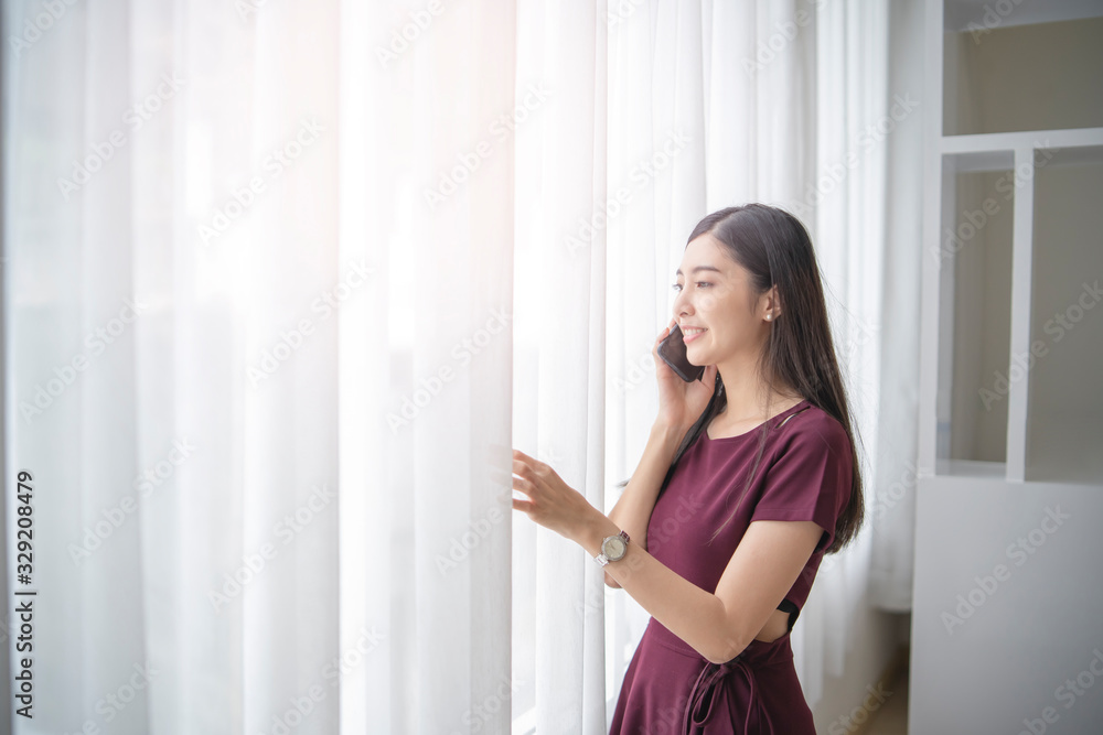 beautiful asian woman smiling while talking or answering on her phone looking out of the window with bright lights shining through long white curtain, and standing up straight touching the curtains.