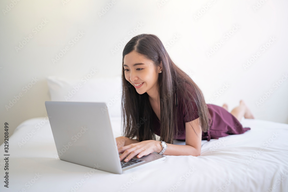 asian beautiful woman lying across on a white bed flat on the belly playing on the laptop typing on the keyboard with both hands while smiling at the laptop screen, long black hair wearing a dress... asian beautiful woman lying across on a white bed flat on the belly playing on the laptop typing on the keyboard with both hands while smiling at the laptop screen, long black hair wearing a dress...