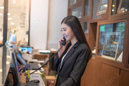 asian woman hotel receptionist holding a wired landline phone against her ear smiling looking down at a computer monitor with a keyboard and a mouse reading information on her desk behind the counter. photo