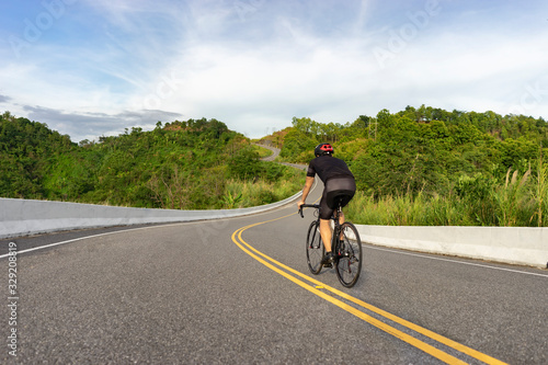back view of a cyclist on top of a mountains winding road, riding a black bicycle down the wearing bike helmet, with grey clouds and blue sky covering the sky and trees of forest in the background.