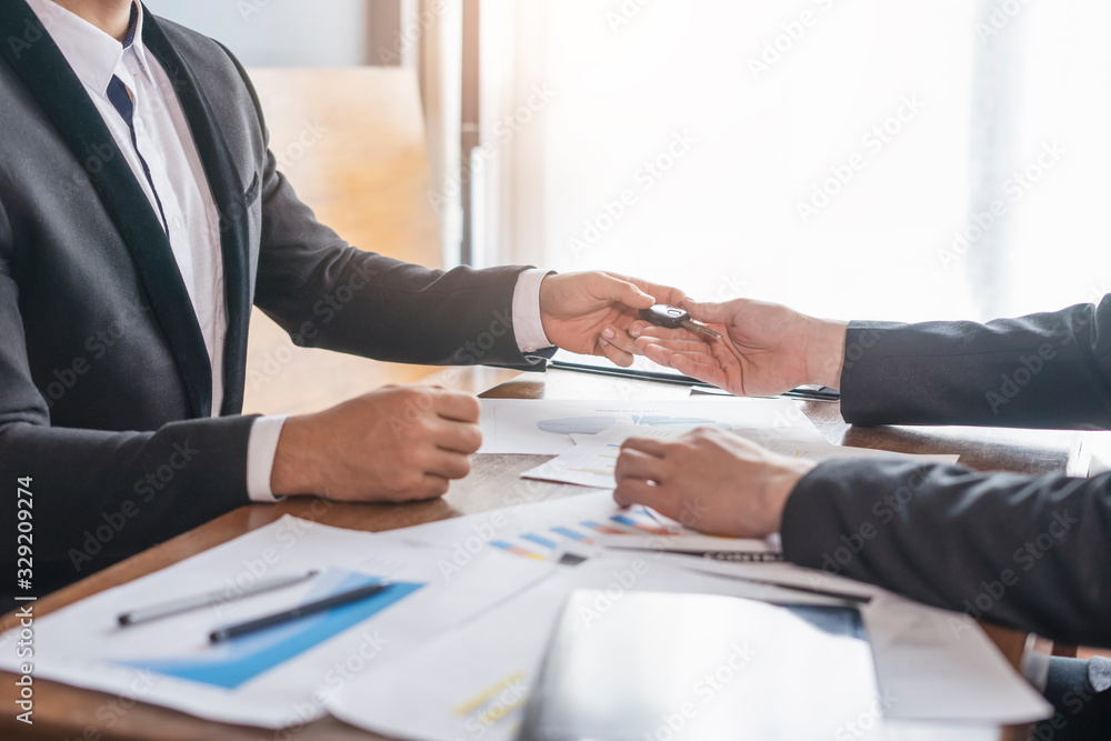 businessman giving car key to his colleague, and reaching over receiving the car key, with work on paper about statistics and data about the business, piling on top of the table with pen and clipboard businessman giving car key to his colleague, and reaching over receiving the car key, with work on paper about statistics and data about the business, piling on top of the table with pen and clipboard