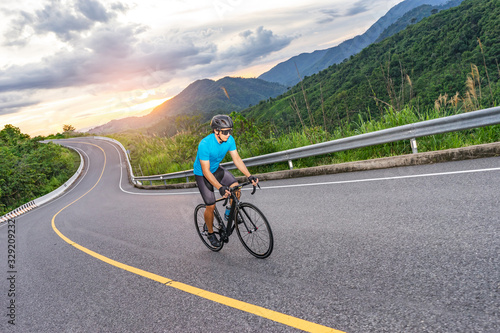 asian male riding on a black bicycle along the winding road up a hill, wearing a cycling blue jersey, crash helmet and goggles, sunset light, grey sky, and forest trees and mountains in the background © Have a nice day 