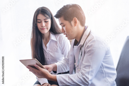 asian male doctor wearing a white scrub lab coat with a stethoscope around his neck smiling and holding a tablet showing health information to a young beautiful asian female patient smartly dressed.