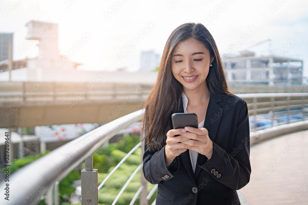 asian business woman holding a smartphone with two hands messaging and smiling, on top of a bridge platform standing next to railing guard with the city buildings and construction in the background. - obrazy, fototapety, plakaty 