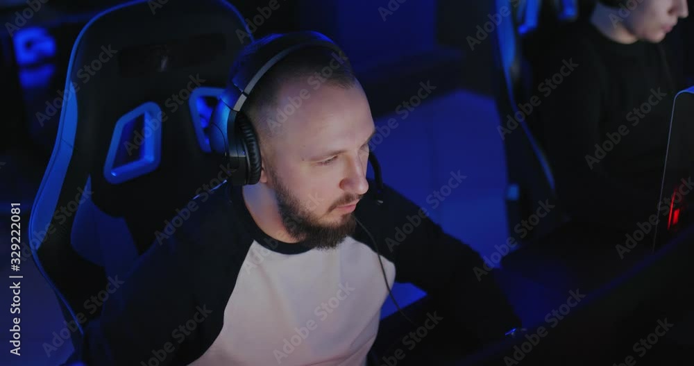 Young man stares intently at the computer monitor, the dark room of the computer club. Man talking to players, an online game. Portrait view. Young man stares intently at the computer monitor, the dark room of the computer club. Man talking to players, an online game. Portrait view.