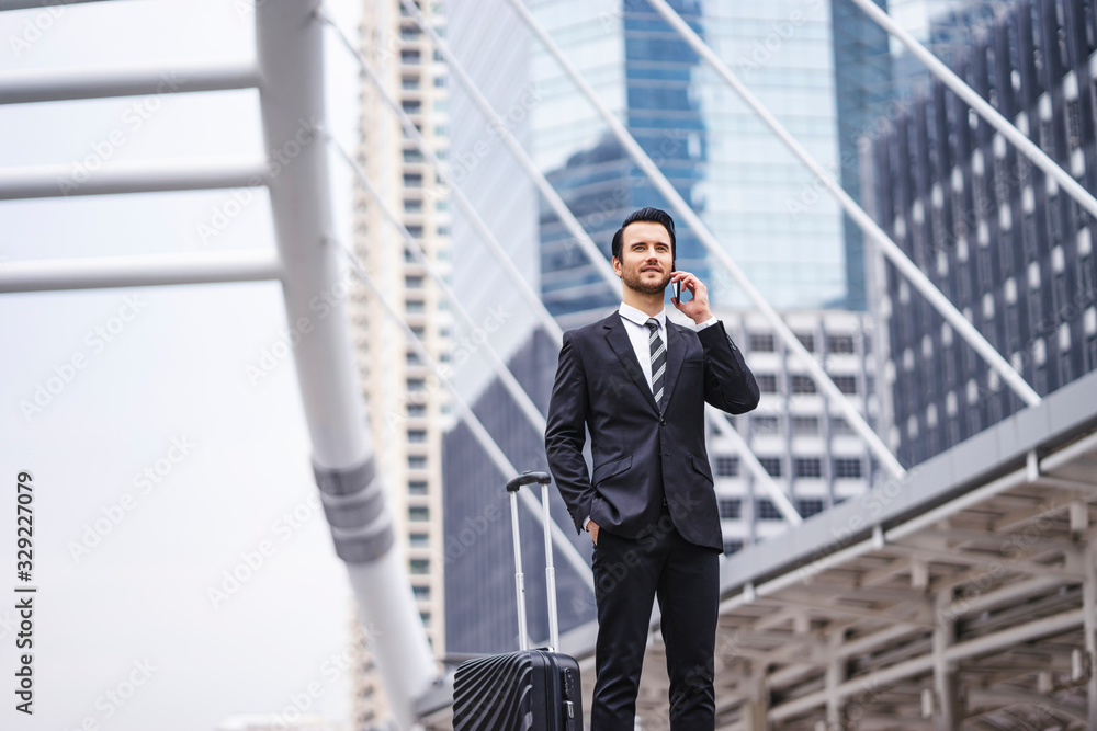 Caucasian businessman using a smart phone talking in a call with his hand in his pocket next to suit case, wearing formally suit and tie with tall city skylines and metro station in the background Caucasian businessman using a smart phone talking in a call with his hand in his pocket next to suit case, wearing formally suit and tie with tall city skylines and metro station in the background