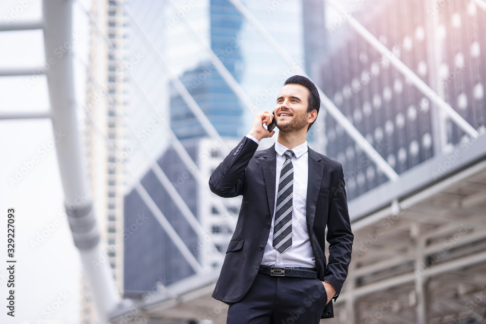 Caucasian businessman standing confidently using a smart phone talking in a call with his hand in his pocket, wearing suit and tie with city skylines and urban architectural structure in background.. Caucasian businessman standing confidently using a smart phone talking in a call with his hand in his pocket, wearing suit and tie with city skylines and urban architectural structure in background..