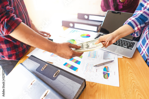 close up of businessman receiving money bills, representing getting paid monthly salary, across the desk from each other with wooden work desk with files, commuter laptop, pen and paper documentations