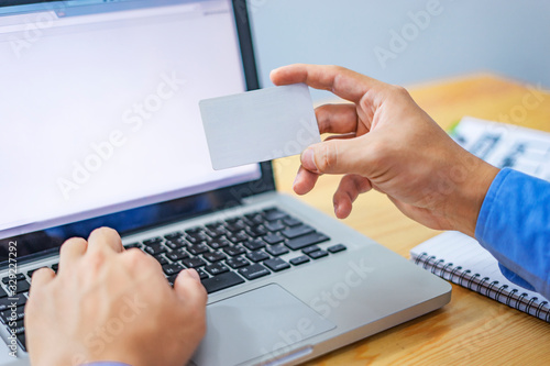 close up of an asian office worker using and typing on his laptop computer and looking at a business card, research and working on his business project, working on his wooden table within his office