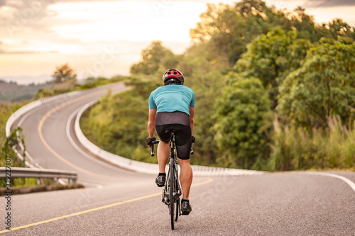 Fototapeta Naklejka Na Ścianę i Meble - back view of a cyclist on top of a mountains winding road, riding a black bicycle down a hill, wearing bike helmet and blue cycling jersey, with grey clouds sunset sky and forest in the background.
