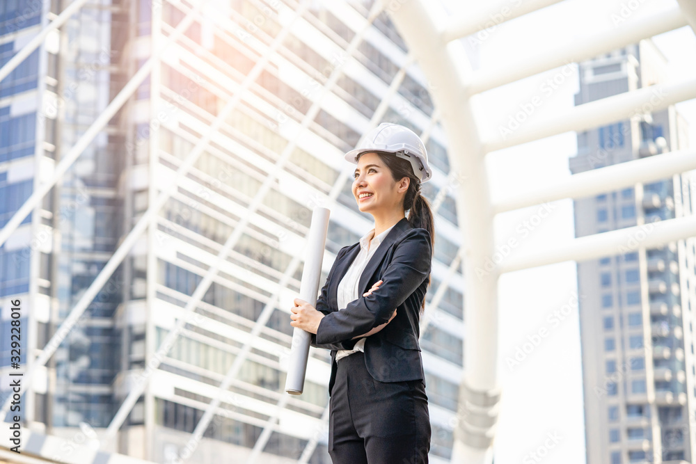 Asian female construction manager with arms cross her chest holding a rolled up blue print, smiling joyfully, formally wearing a suit and a white hardhat for safety, within an urban city district Asian female construction manager with arms cross her chest holding a rolled up blue print, smiling joyfully, formally wearing a suit and a white hardhat for safety, within an urban city district