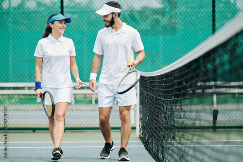 Cheerful couple walking along the net and enjoying discussing game of tennis they just had