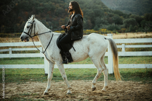 Handsome bearded man riding horse at farm. Beautiful horse with man rider trotting on autumnal field. Equestrian and animal love concept. Rider on gray arabian horse in the field.