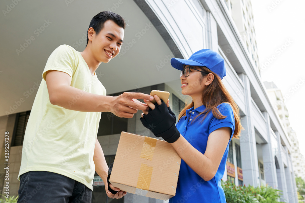 Smiling young delivery woman giving package and asking customer to sign document on her smartphone