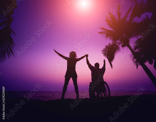 Disabled man in a wheelchair with his wife on the beach. Silhouettes at sunset