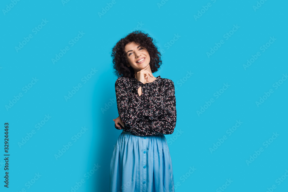 Caucasian lady with curly hair is touching her chin while wearing a beautiful dress and posing on a blue wall Caucasian lady with curly hair is touching her chin while wearing a beautiful dress and posing on a blue wall