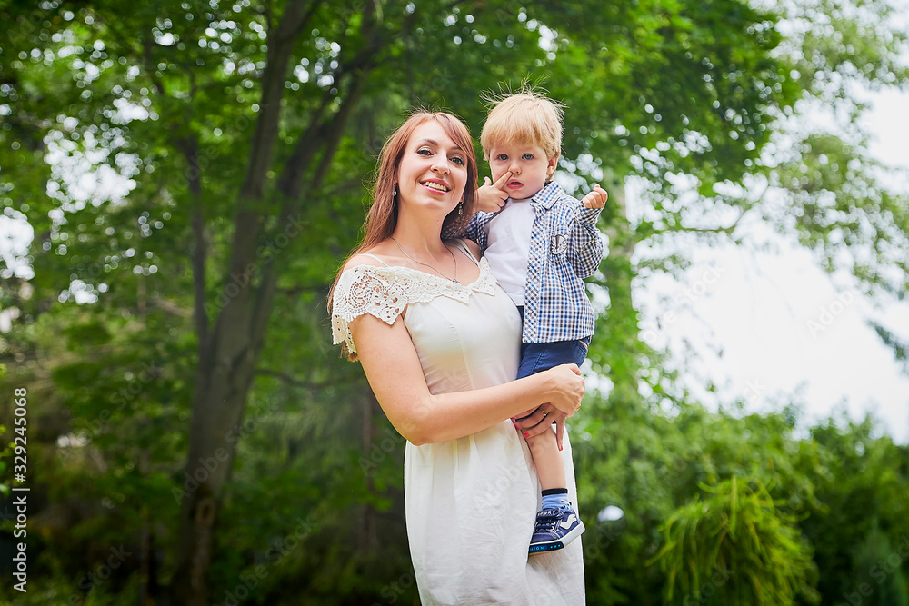 Fototapeta premium Beautiful young woman in a white dress and long hair with a cute little baby blonde boy at the green garden in a day