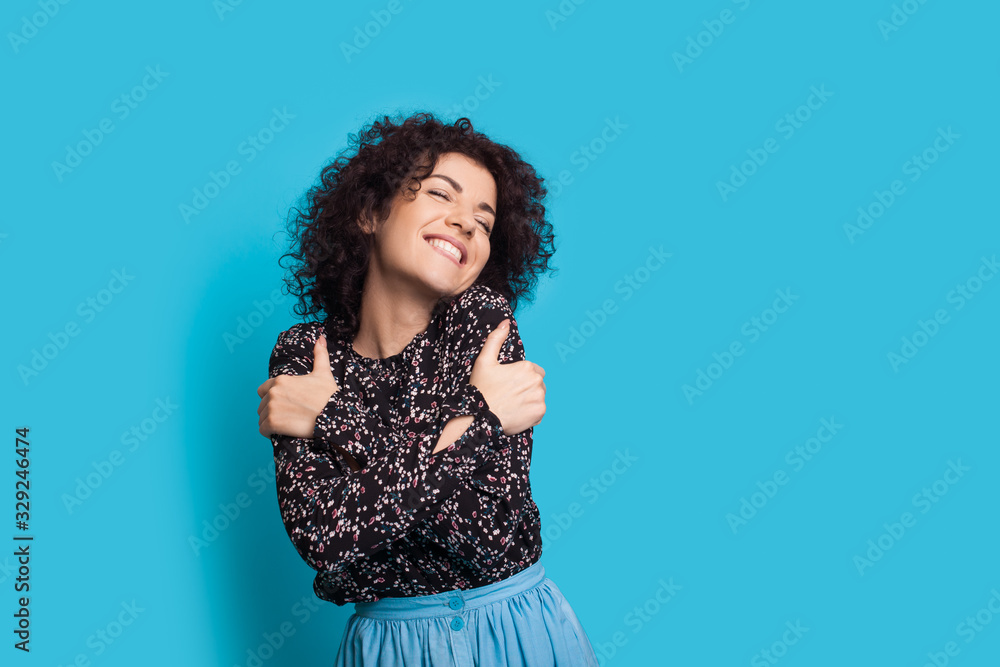 Cheerful caucasian lady with curly hair embracing herself on blue background while smiling