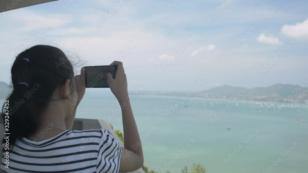 Rear view of pretty girl holding mobile phone while taking picture on view point tower with beautiful scenery of the bay with cloudy blue sky in phuket. Rear view of pretty girl holding mobile phone while taking picture on view point tower with beautiful scenery of the bay with cloudy blue sky in phuket.