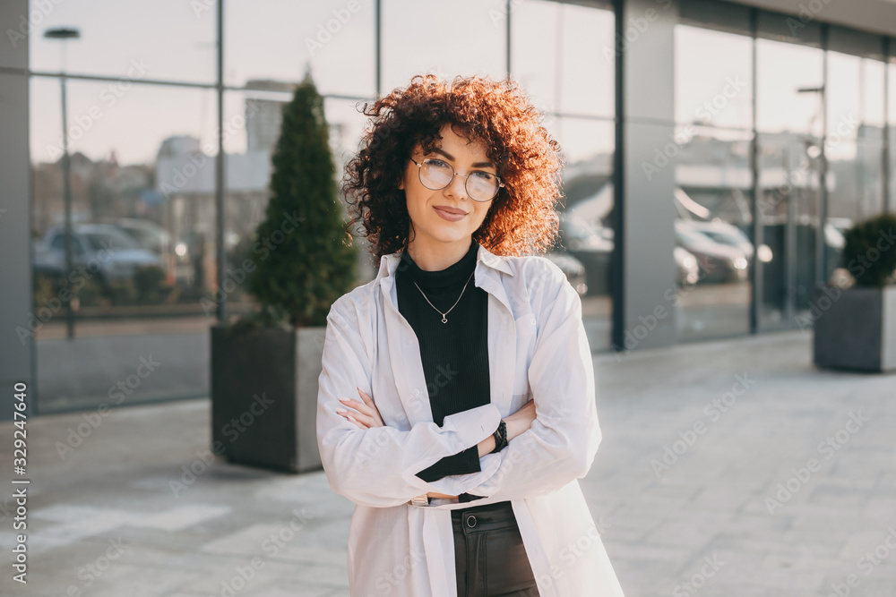 Charming caucasian woman with curly hair is looking through eyeglasses while posing outside with crossed hands Charming caucasian woman with curly hair is looking through eyeglasses while posing outside with crossed hands