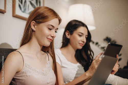 Two concentrated caucasian lady are spending their time sitting on a sofa and surfing on internet using a tablet and laptop