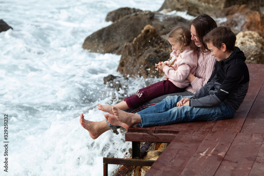 Happy mom, eldest son and little daughter sit barefoot on a wooden pier and laugh on the background of the sea and rocks,
