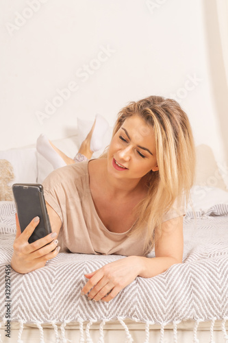 A girl in pajamas is lying on a bed in her apartment with a phone. She is surfing in social networks and smiling. Soft pink creamy colors of the picture. Vertical picture