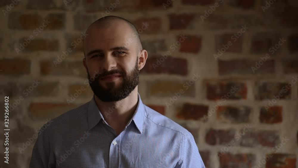 A bald office worker turns his head and smiles with the brick wall on the background. A man wears two silver earrings and a blue shirt. A bald office worker turns his head and smiles with the brick wall on the background. A man wears two silver earrings and a blue shirt.