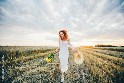 Beautiful young curly red-haired woman in field at the sunset. Girl having fun.