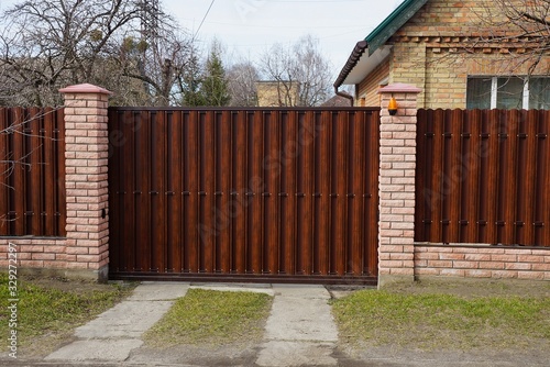 brown closed private gate and part of a fence of wooden boards and bricks on a rural street in green grass