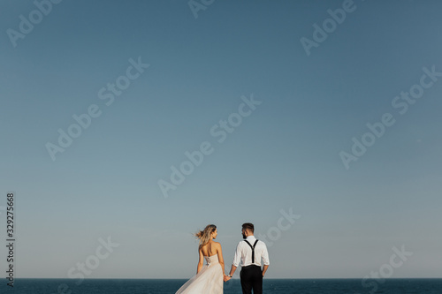 Back view of beautiful wedding couple bride and groom at wedding day outdoors at sea. Loving emotional man and woman at marriage day at blue sky and sea background. Loving newlyweds hold hands.