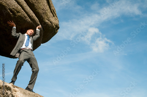Strong businessman struggling to lift massive boulder into blue sky copy space