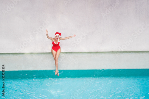 Enjoying christmas holidays and vacation. Top view of excited young woman in red swimsuit and santa claus hat rising hands up near swimming pool.