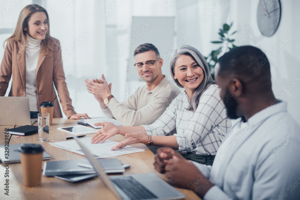 Fototapeta premium smiling asian woman pointing with hands at folder and looking at african american colleague in creative agency