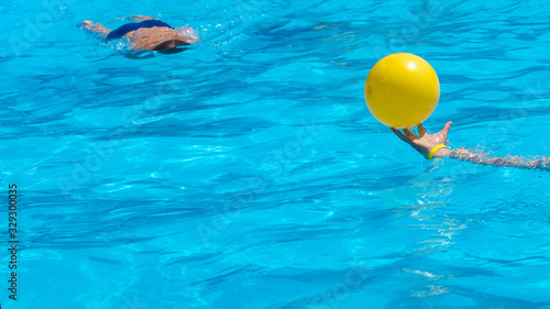Background In the pool  blurred figure of a diving boy and a hand holding a yellow ball in a refreshing blue pool
