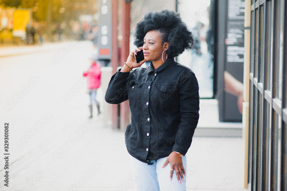 Attractive happy dark-skinned model with Afro hairstyle and nose-ring, posing outdoors against urban background during her morning walk, looking down with shy smile showing her white teeth. Flare sun