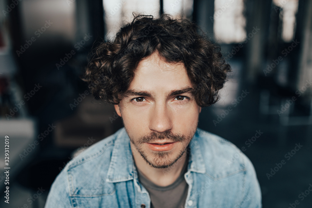 Close up portrait of handsome guy with curly hair in blue jeans shirt looking to camera Close up portrait of handsome guy with curly hair in blue jeans shirt looking to camera