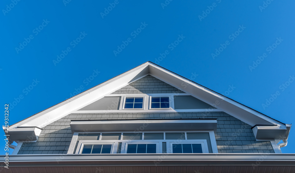 Decorative gable with white gutters and soffit, gray shingle sidings, bump-out windows framed with thick white frame with blue sky on a new construction American house