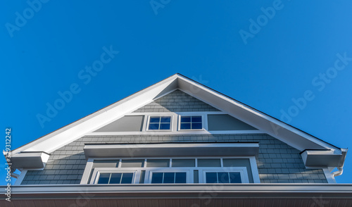 Decorative gable with white gutters and soffit, gray shingle sidings, bump-out windows framed with thick white frame with blue sky on a new construction American house © tamas