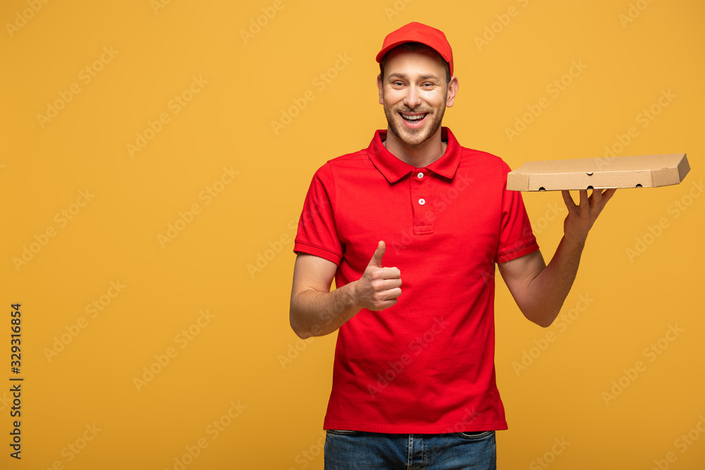happy delivery man in red uniform holding pizza box and showing thumb up isolated on yellow