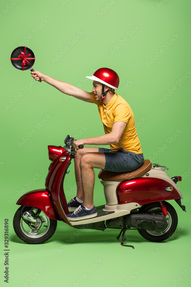 side view of delivery man in yellow uniform holding frying pan with bow on scooter on green