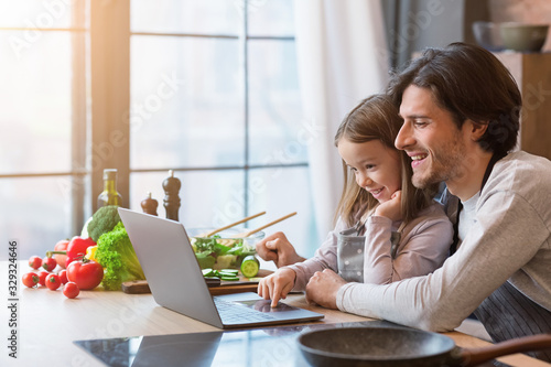 Little girl and her dad using laptop at kitchen, looking recipe