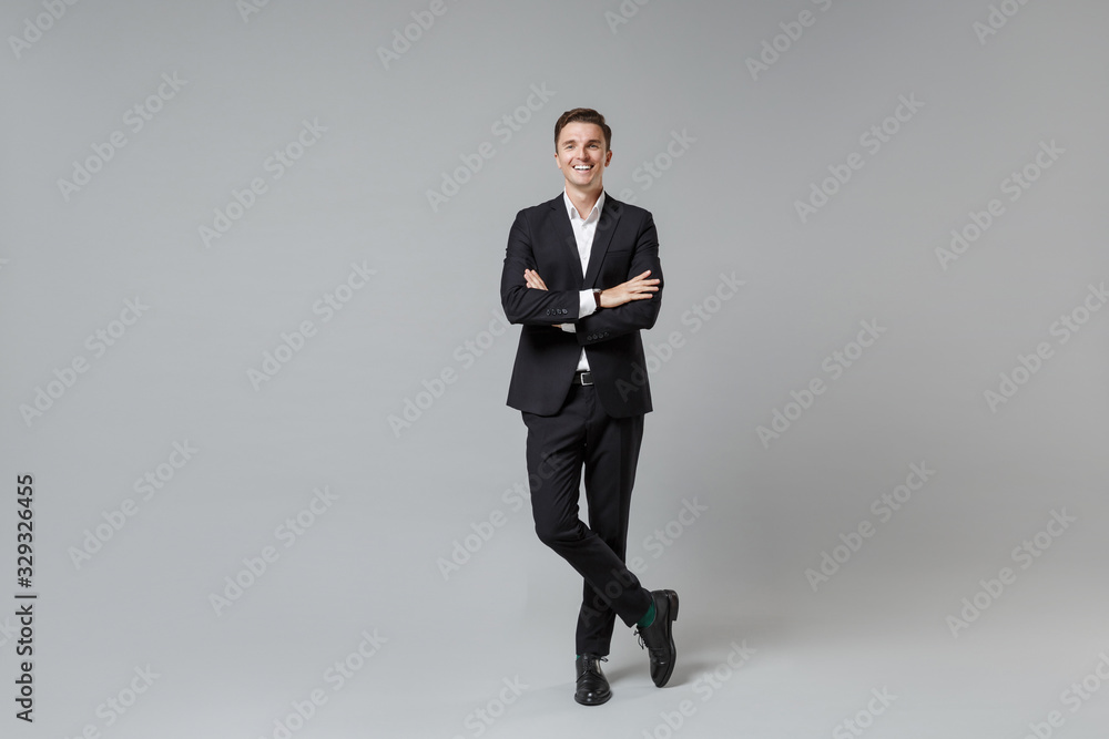 Cheerful young business man in classic black suit shirt posing isolated on grey wall background studio portrait. Achievement career wealth business concept. Mock up copy space. Holding hands crossed. Cheerful young business man in classic black suit shirt posing isolated on grey wall background studio portrait. Achievement career wealth business concept. Mock up copy space. Holding hands crossed.